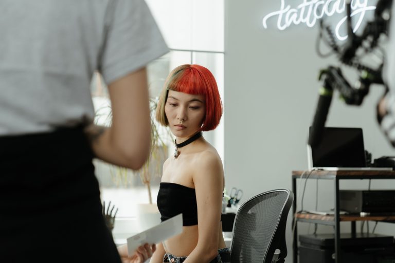 A woman with red hair sits in a tattoo studio while another person shows her a sheet of paper