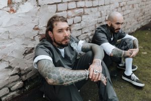 Two men in matching gray uniforms sit against a weathered brick wall, their forearms covered in tattoos.