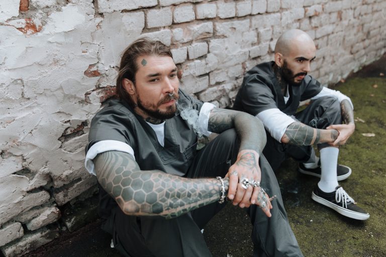 Two men in matching gray uniforms sit against a weathered brick wall, their forearms covered in tattoos.